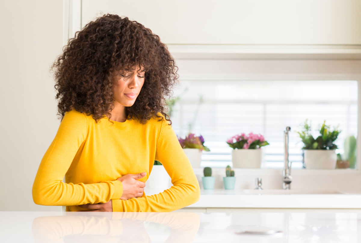 Afro-American woman wearing a yellow sweater in the kitchen with hand on the stomach because nausea, painful illness feeling unwell.
