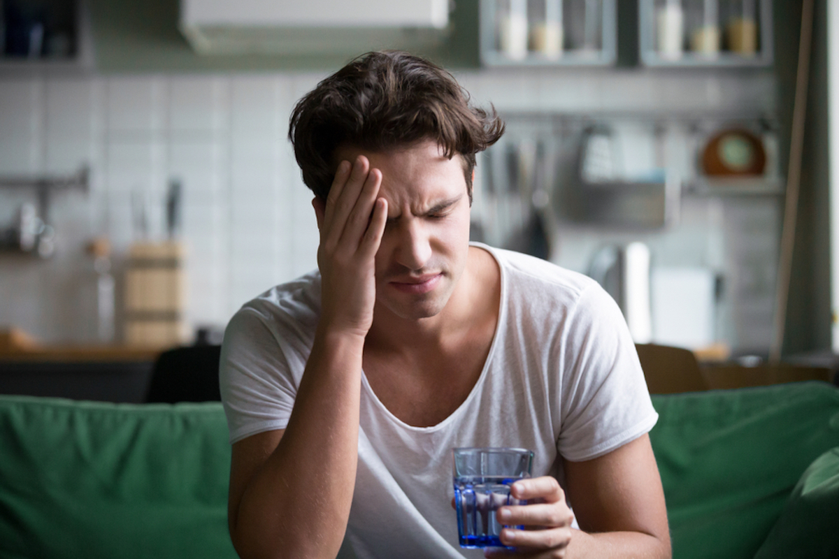 Young man suffering from strong headache or migraine sitting with a glass of water in the kitchen, a millennial man suffering intoxication and pain touching aching head, the morning after hangover concept