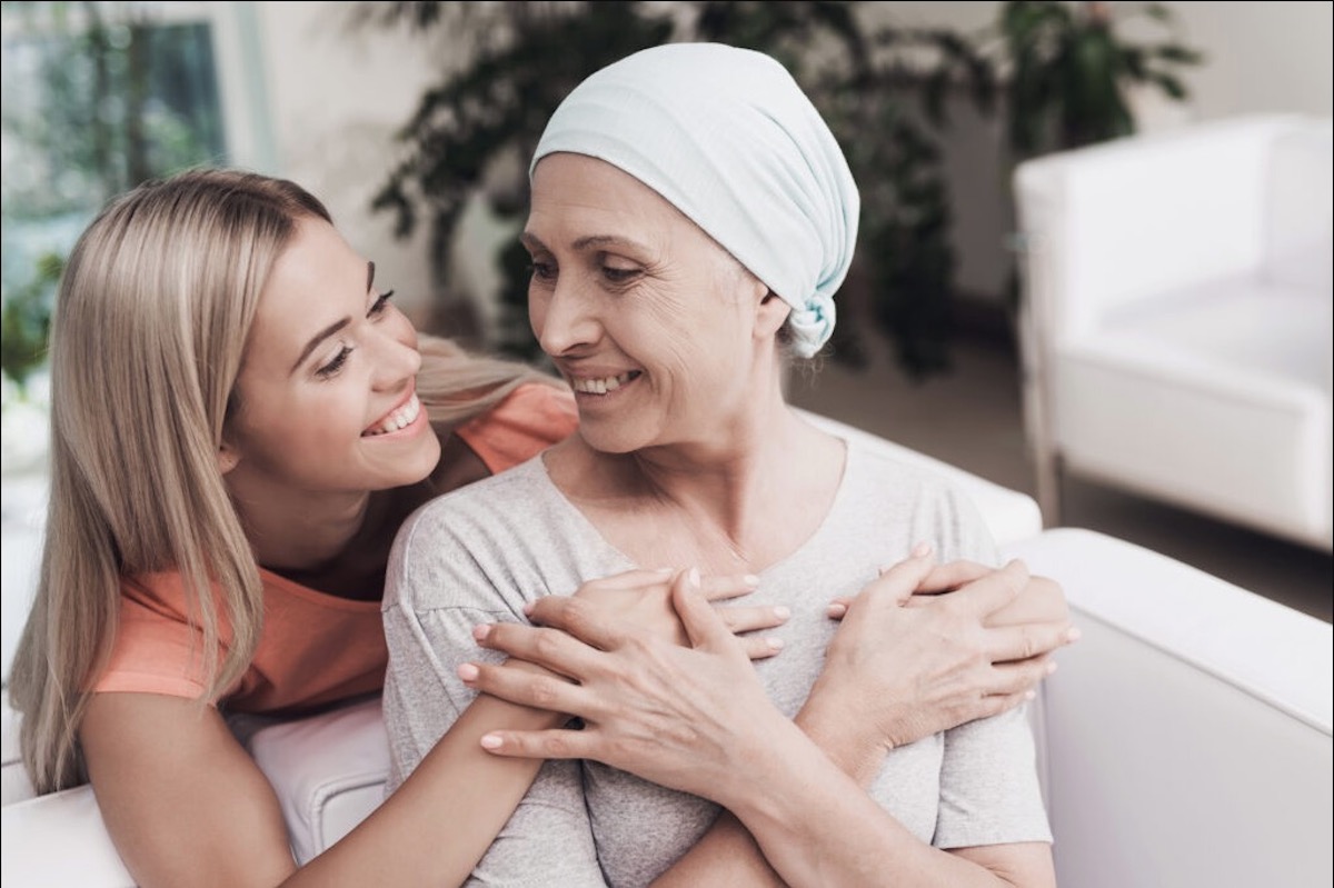 One younger Caucasian woman with blond hair and an orange shirt is hugging an older Caucasian woman with a light bandana on her head. The older woman probably has cancer.