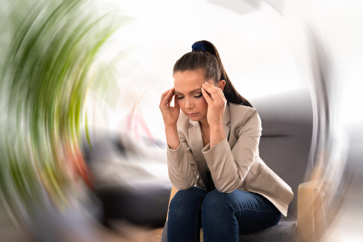 Caucasian woman with a high ponytail sitting on a gray/wooden chair while bending forward. She has her fingers to her scalp and eyes closed. She possibly suffers from vertigo.